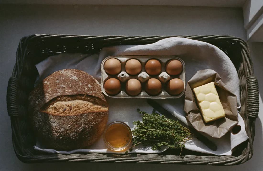 An open wicker hamper with sourdough, eggs, honey and butter on a scrubbed oak table