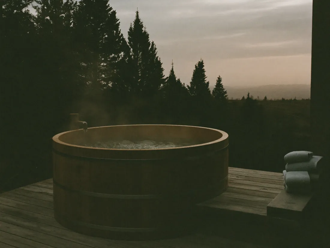 A round cedar hot tub steaming on a timber deck at dusk with pines behind