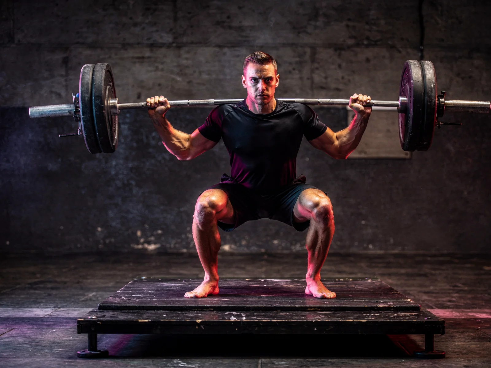 An athlete under a loaded barbell in magenta and cyan light